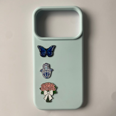 Pinkgul.no light blue phone case decorated with three metal stickers: a blue butterfly, a hamsa hand, and a flower bouquet.